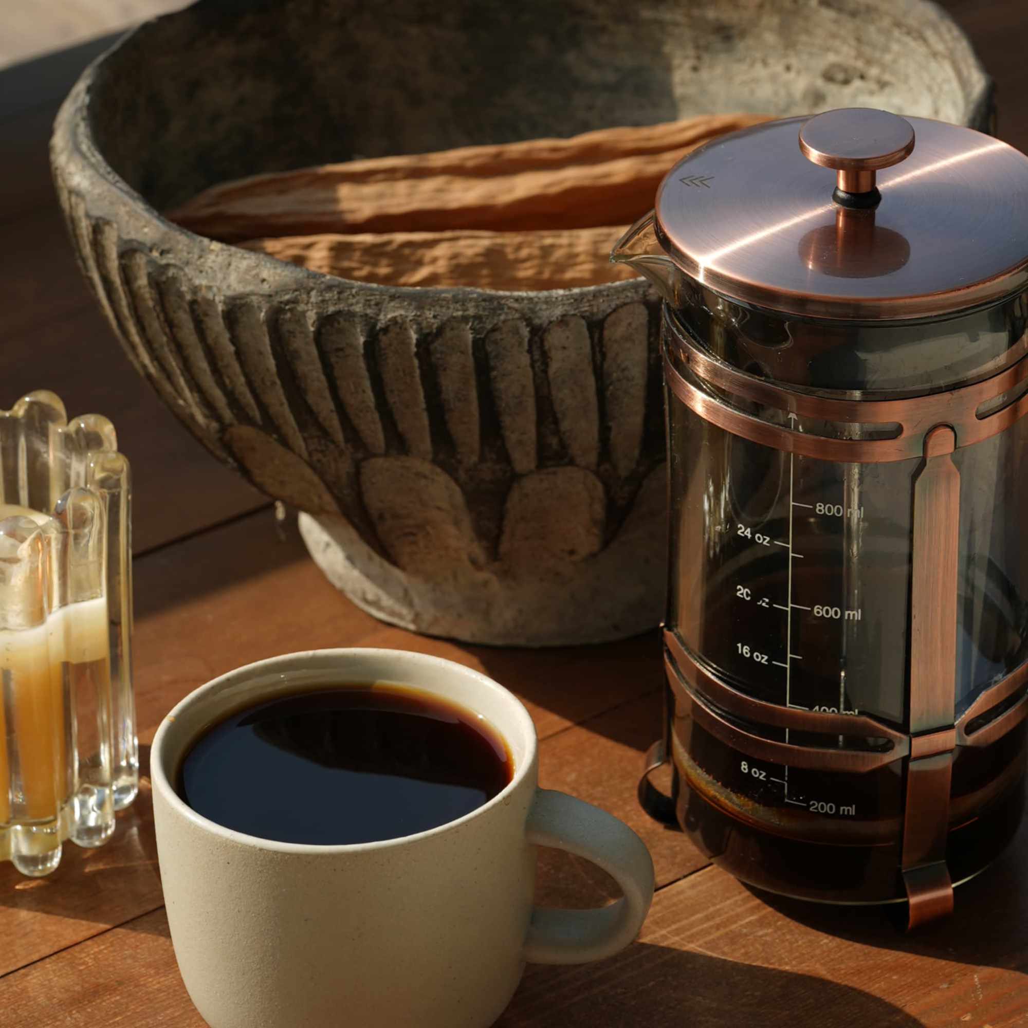 Prensa francesa de cobre y taza de café AM sobre madera, junto a un molcajete de piedra
