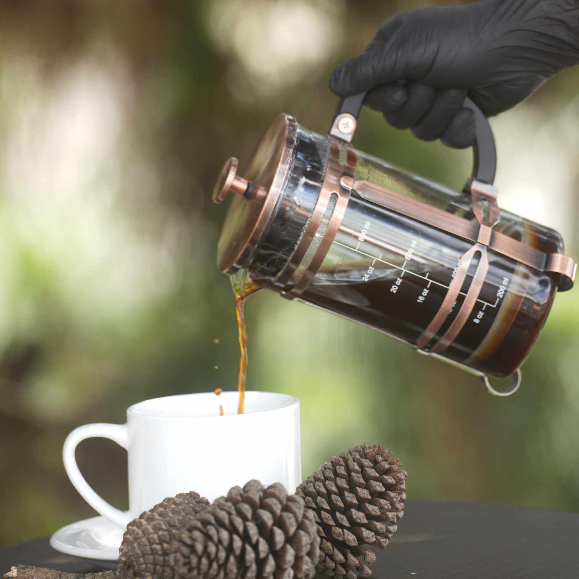 Mano sirviendo café desde una prensa francesa de cobre junto a piñas de pino