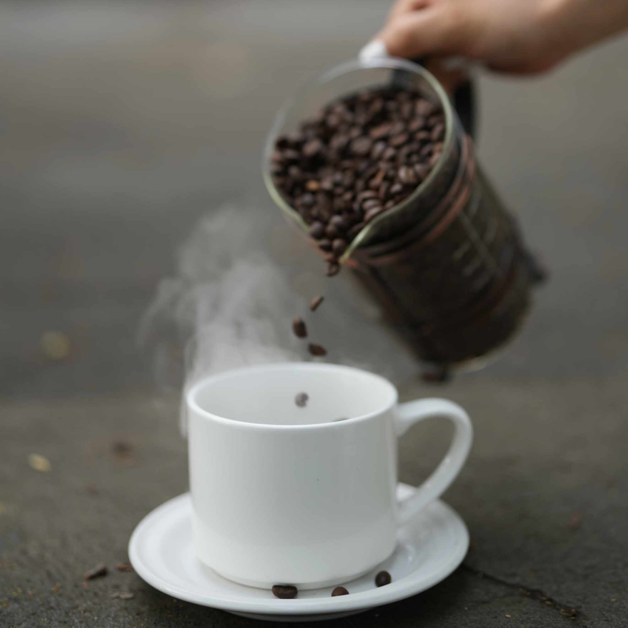 Granos de café cayendo desde una jarra hacia una taza blanca humeante