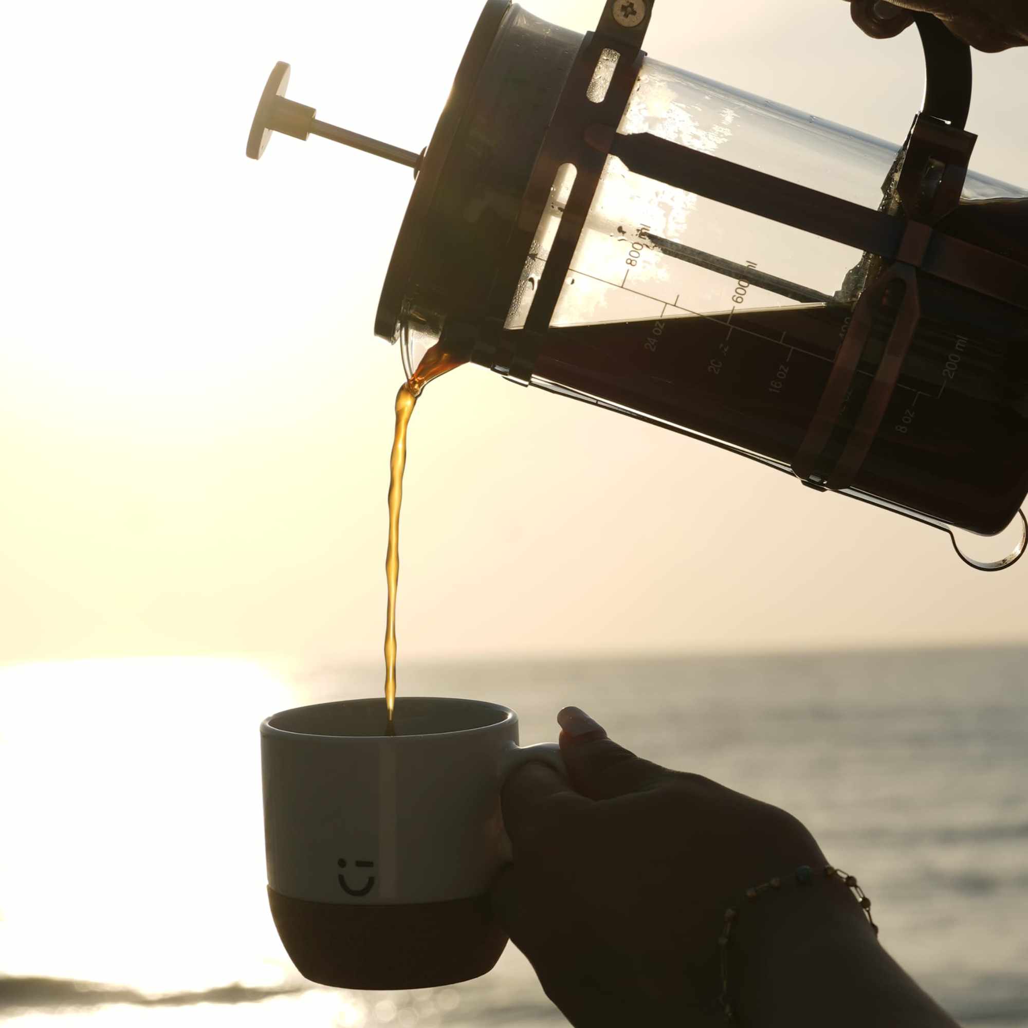 Silueta sirviendo café desde una prensa francesa frente al mar al amanecer