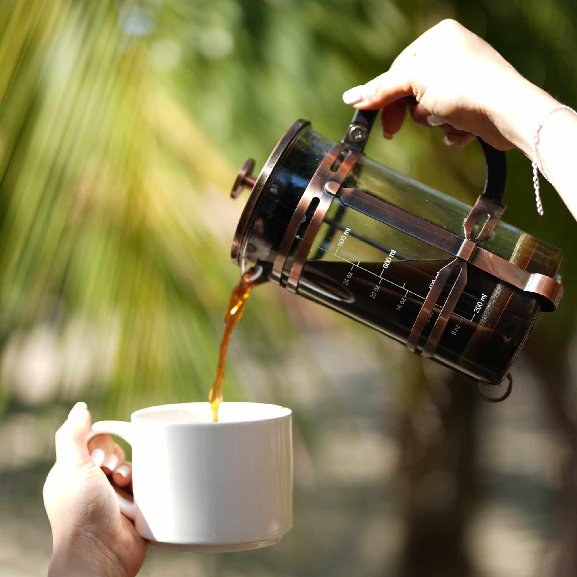 Persona sirviendo café desde una prensa francesa de cobre entre vegetación tropical